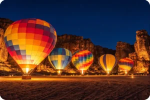Illuminated hot air balloons beneath sandstone cliffs against a dark blue sky