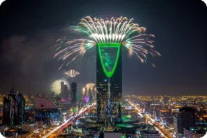 Fireworks above a green-lit tower overlooking a city skyline at night.