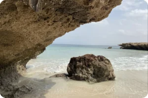 Coastal view beneath a weathered rock overhang, with pale sand and calm turquoise water.
